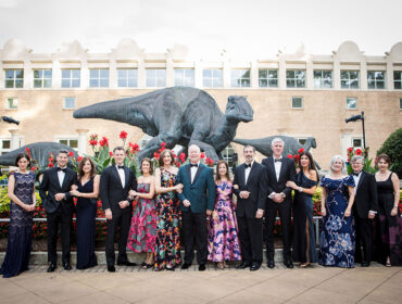 Group in formal wear poses in front of dinosaur statues and a brick building.