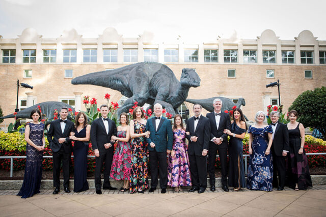 Group in formal wear poses in front of dinosaur statues and a brick building.