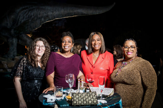 Four women smiling at a table with drinks and clutches. A dinosaur sculpture is in the background.