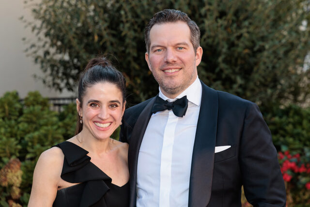 Couple in formal wear smiling outdoors with greenery in the background.