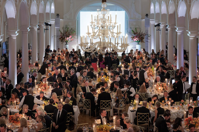Grand ballroom with long tables set for a formal dinner, ornate chandelier, and white columns.