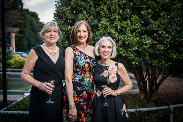 Three women in formal dresses holding champagne glasses outdoors.