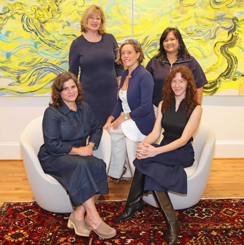 Five women posing in front of a large abstract yellow and blue painting.