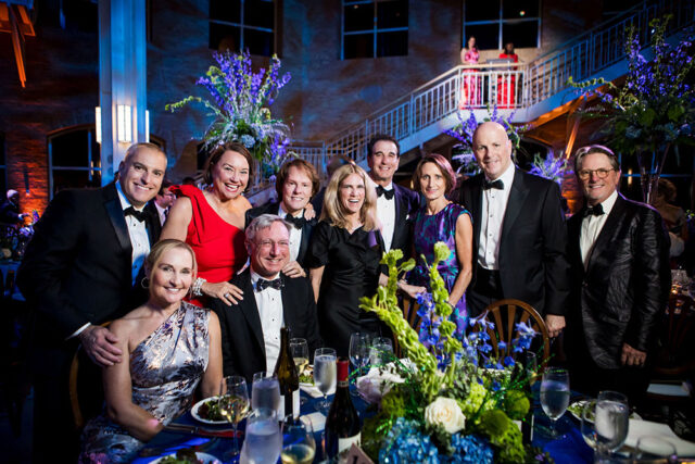 Group of people in formal attire posing for a photo at a formal event with floral arrangements.
