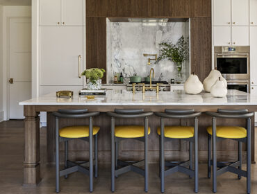 Kitchen island with marble countertop, yellow bar stools, and wood cabinetry. Marble backsplash