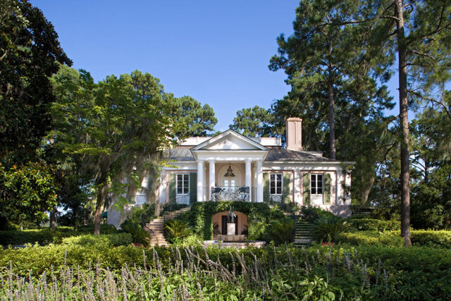 White house with columns and green shutters, surrounded by lush trees and gardens.