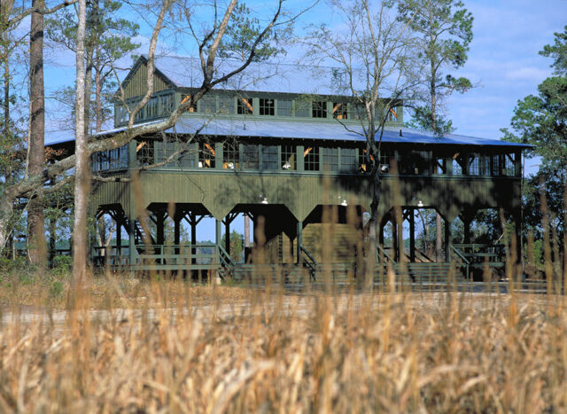 Green wooden building with a metal roof, surrounded by trees and dry grass.