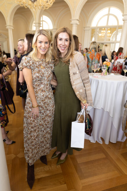 Two women smiling at a party. One wears a floral dress, the other a green dress and textured jacket.