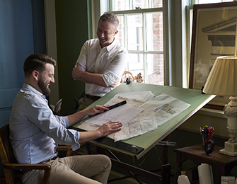 Two men reviewing architectural drawings at a drafting table in a room with green walls and a large