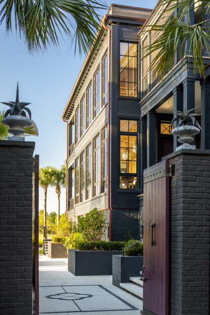 Dark gray brick gateposts with urns flank a dark wood gate leading to a dark gray house with many