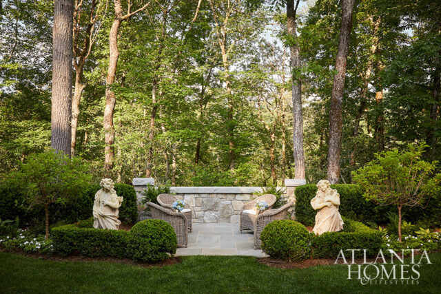 Stone patio with wicker chairs and statues surrounded by lush greenery and trees.