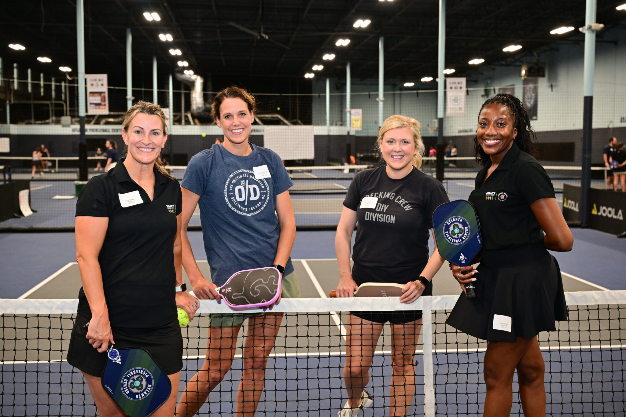 Four women in athletic wear stand behind a pickleball net, holding paddles.