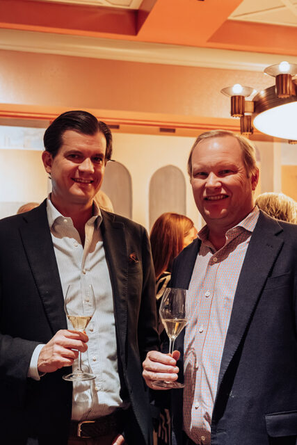 Two men in suits holding champagne flutes, smiling.