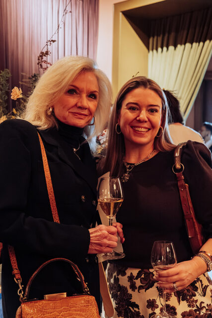 Two women smiling, holding wine glasses. One has blonde hair, the other has brown hair.
