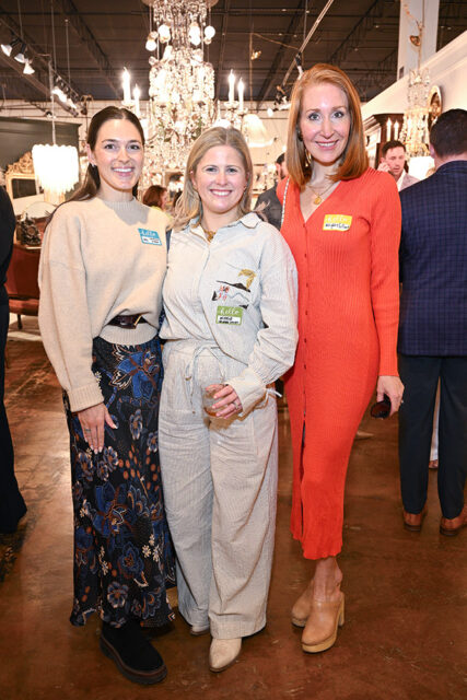 Three women pose for a photo in a store with chandeliers and furniture in the background. The woman on the left wears a tan sweater and floral skirt. The woman in the middle wears a striped jumpsuit and holds a drink. The woman on the right wears an orange ribbed dress.