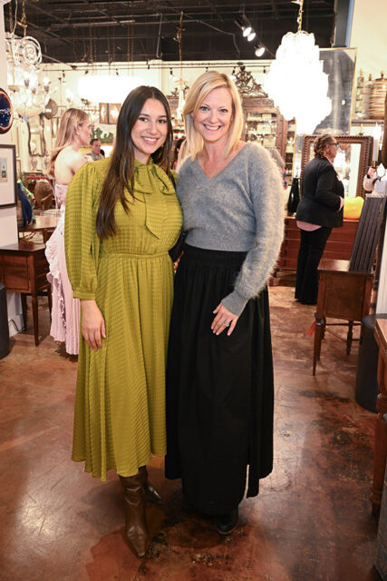 Two women smiling at the camera in an antique store. The woman on the left wears a textured olive green dress and brown boots. The woman on the right wears a gray fuzzy sweater and a long black skirt.