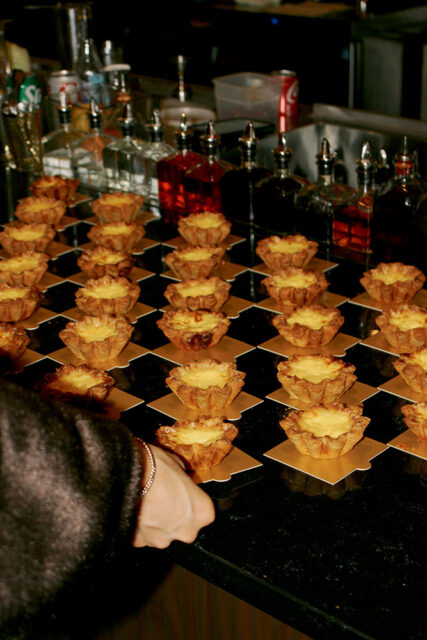 Rows of golden custard tarts with fluted pastry shells arranged on a black and gold checkered surface, with bottles of liquor blurred in the background.