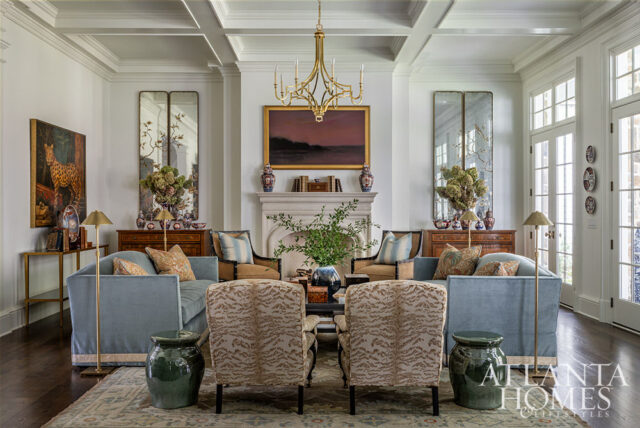 Light blue sofas and patterned armchairs arranged around a stone fireplace with a landscape painting above. Coffered ceiling and tall mirrors flank the fireplace.