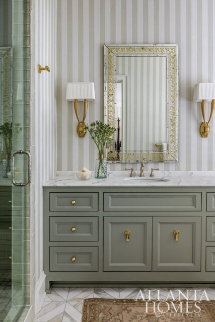 Sage green vanity with brass hardware, marble countertop, and striped wallpaper. Features a Greek key mirror and brass sconces.