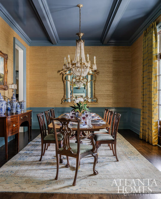 Dining room with grasscloth wallpaper, blue wainscoting, coffered ceiling, ornate chandelier, and a wooden dining table set for a meal.