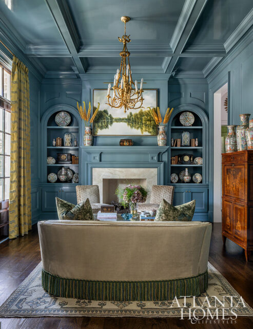 Blue paneled room with coffered ceiling, fireplace, built-in bookshelves, and a curved sofa with green patterned pillows.