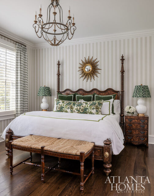 Bedroom with a dark wood four-poster bed, white bedding, green floral pillows, striped wallpaper, a sunburst mirror, and a woven bench.