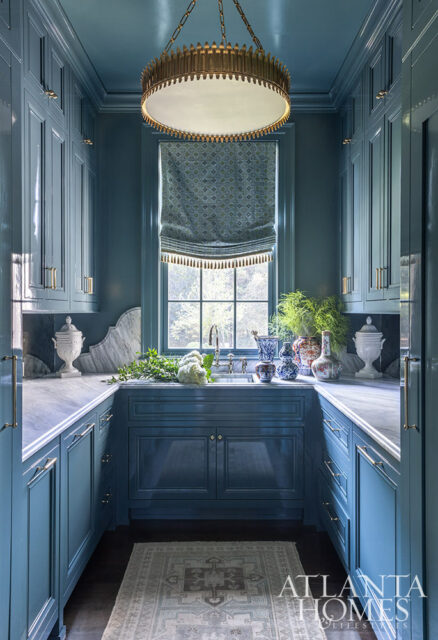 Blue kitchen with marble countertops, brass hardware, and a large ornate chandelier. A patterned roman shade covers the window above the sink.