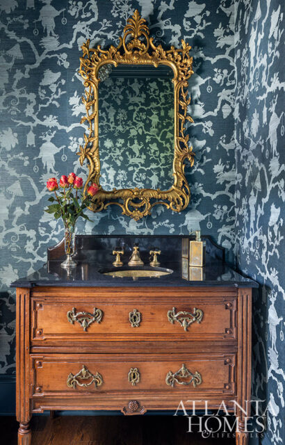 Powder room with dark blue chinoiserie wallpaper, a vintage wooden vanity with ornate brass hardware, a black granite countertop, a brass sink and faucet, and a gold ornate mirror.