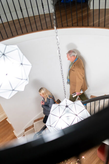 A woman in a grey jacket and a man in a tan coat walk up a curved staircase with white walls, black railings, and geometric pendant lights.
