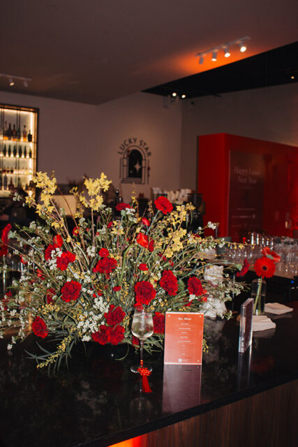 A large floral arrangement of red and yellow flowers sits on a black countertop in front of a bar with bottles on shelves. A sign reads 'Bar Menu'.