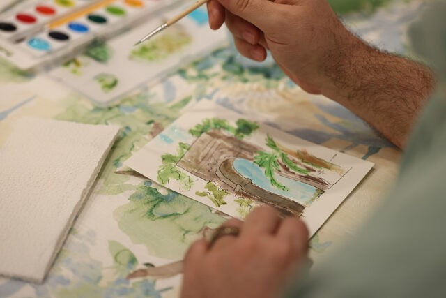 Close-up of a person's hands painting a watercolor landscape featuring a bridge and water, with a palette of watercolors and a brush nearby.