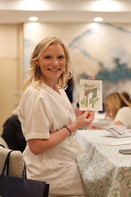 Woman with blonde wavy hair wearing a white dress, holding up a small watercolor painting of an arched entryway with greenery.