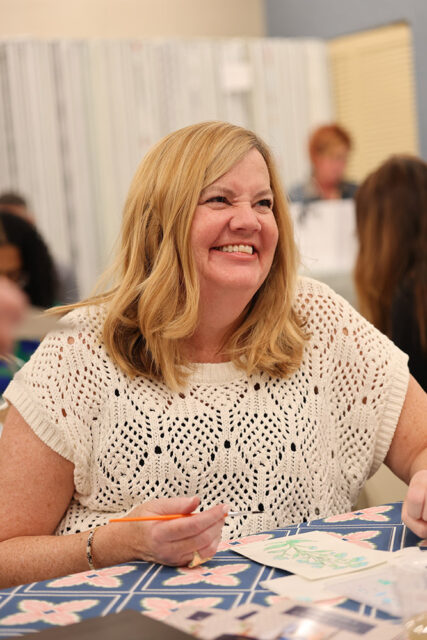 Woman with blonde hair smiling and holding a paintbrush, wearing a cream-colored crocheted top.