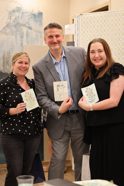 Three people smiling and holding up small artworks. The woman on the left holds a floral illustration. The man in the middle holds a sketch of two figures. The woman on the right holds an illustration of an orange-flowered branch.