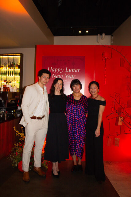 Four people stand in front of a red wall with "Happy Lunar New Year" text. A man in a cream suit stands on the left, followed by three women in dresses.