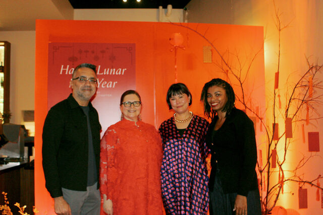 Four people stand in front of an orange backdrop with Lunar New Year decorations. A man in a dark shirt and grey pants smiles. A woman in a bright orange lace dress smiles. A woman in a patterned dress and gold necklace smiles. A woman in a black cardigan smiles.