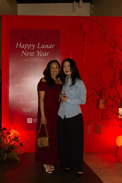 Two women stand in front of a red backdrop with "Happy Lunar New Year" text and sponsor logos. One woman wears a maroon off-the-shoulder dress and the other wears a blue striped shirt and black pants.
