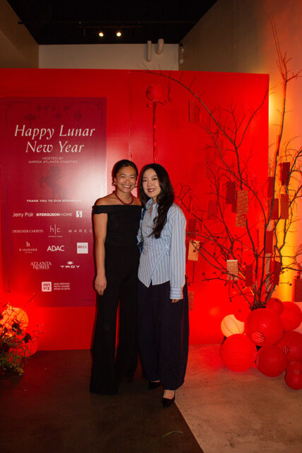 Two women pose in front of a red backdrop with a "Happy Lunar New Year" sign and a decorative tree with hanging ornaments.