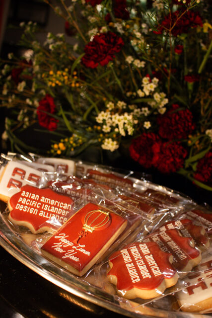 Red cookies decorated with 'Happy Lunar New Year' text and a gold lantern, and cookies with 'asian american pacific islander design alliance' text, arranged on a silver platter with a floral arrangement in the background.