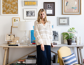 Woman in a cream and white striped sweater and navy skirt stands in front of a console table displaying artwork, books, and decorative objects.
