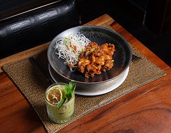 A dark-rimmed bowl of glazed chicken pieces topped with shredded white vegetables sits on a woven placemat next to a glass of green cocktail garnished with a lemon slice and mint leaves.