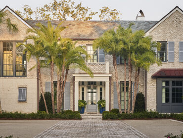 Brick home with gray shutters and a copper roof bay window, featuring a covered entryway with palm trees and manicured landscaping.