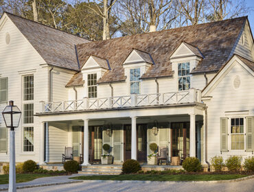 White clapboard house with a cedar shake roof, dormer windows, and a wraparound porch with white railings and columns.