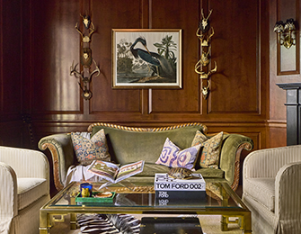 Living room with dark wood paneled walls, a green velvet sofa with patterned pillows, and two cream upholstered armchairs around a brass and glass coffee table.