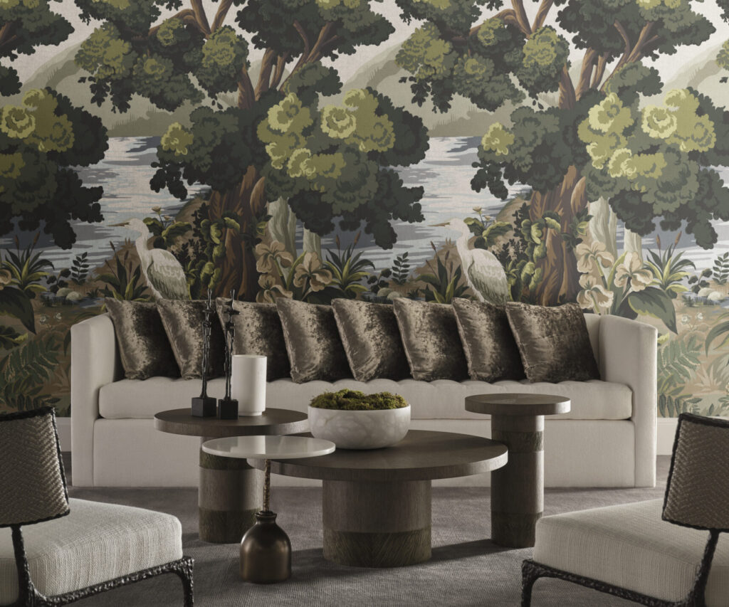Living room with a cream sofa adorned with velvet pillows, set against a scenic wallpaper featuring trees, water, and a heron. Round wooden coffee tables and textured chairs complete the space.