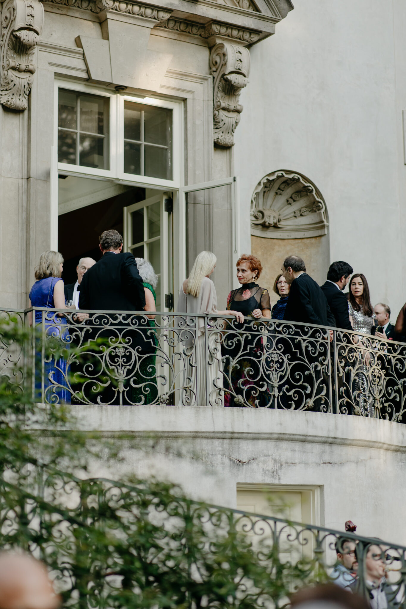 People in formal attire gathered on a balcony with ornate iron railings and classical architectural details.