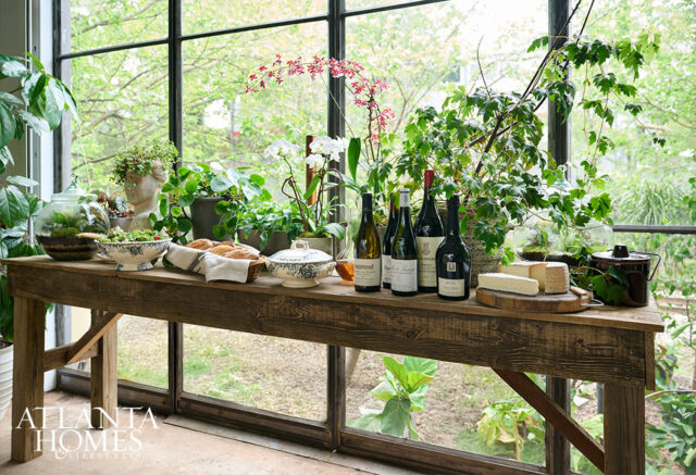 Wooden table with wine bottles, cheese, bread, and plants in front of a large window.