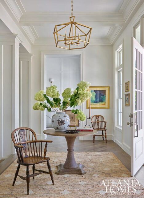 Entryway with round pedestal table, ginger jar with hydrangeas, and wooden chairs.