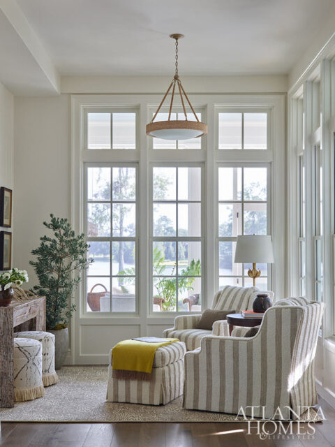 Living room with striped armchairs, ottoman with yellow throw, and large windows overlooking water