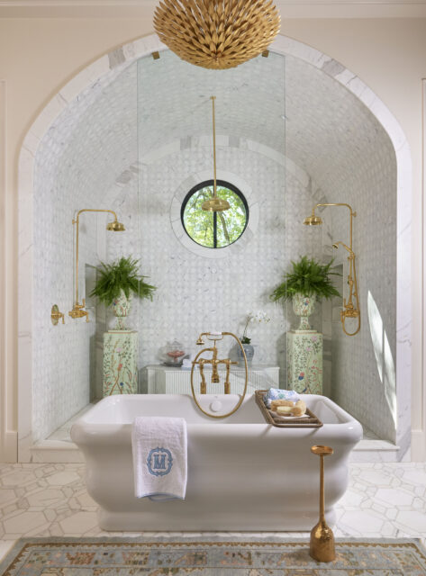 White freestanding bathtub with gold fixtures, arched marble shower, and ornate chandelier.